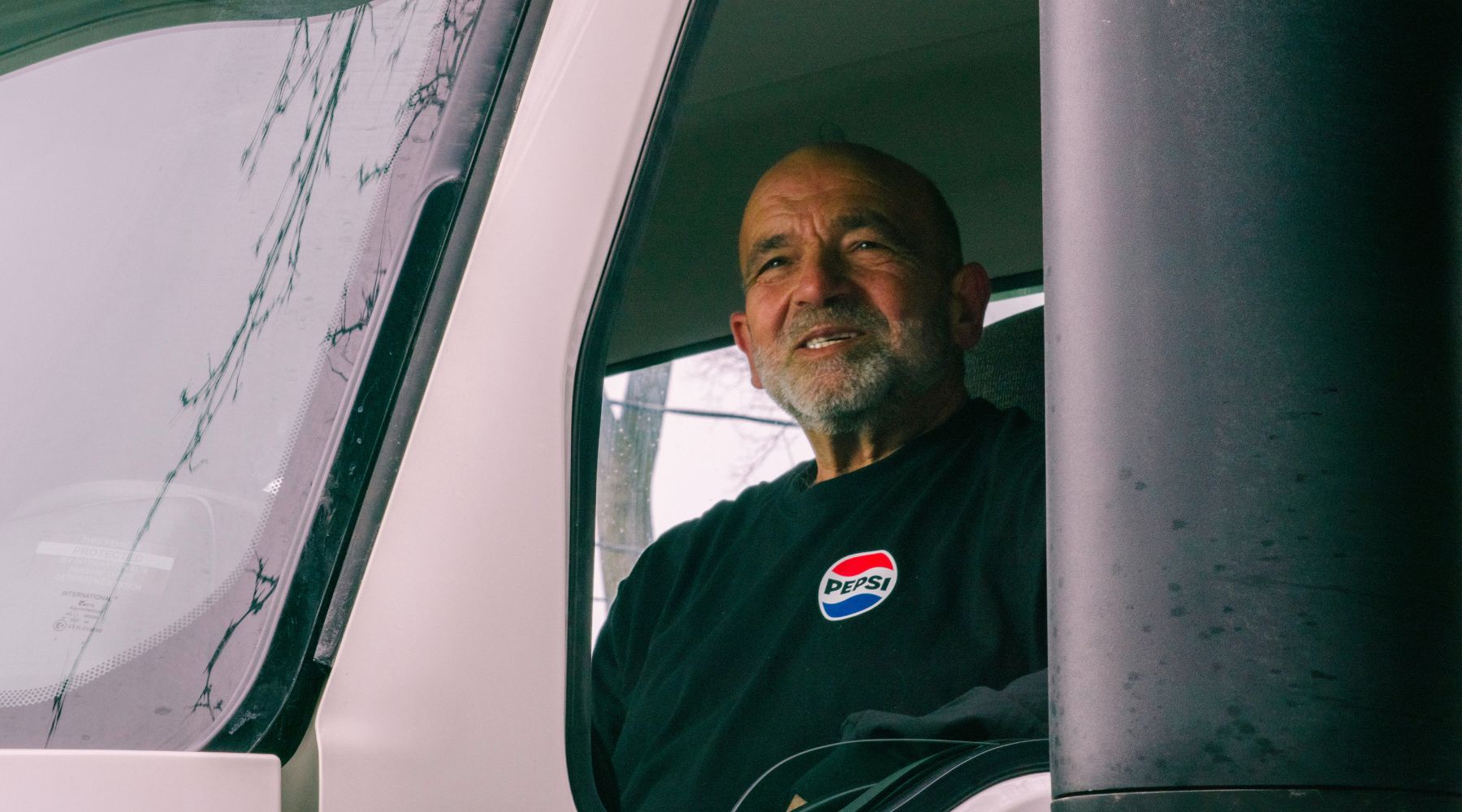 PepsiCo Canada employee Bob, wearing a black Pepsi-branded shirt is seated in the driver’s seat of a vehicle, viewed through the open window. The interior of the vehicle and part of the side mirror are visible, along with raindrops on the window frame.