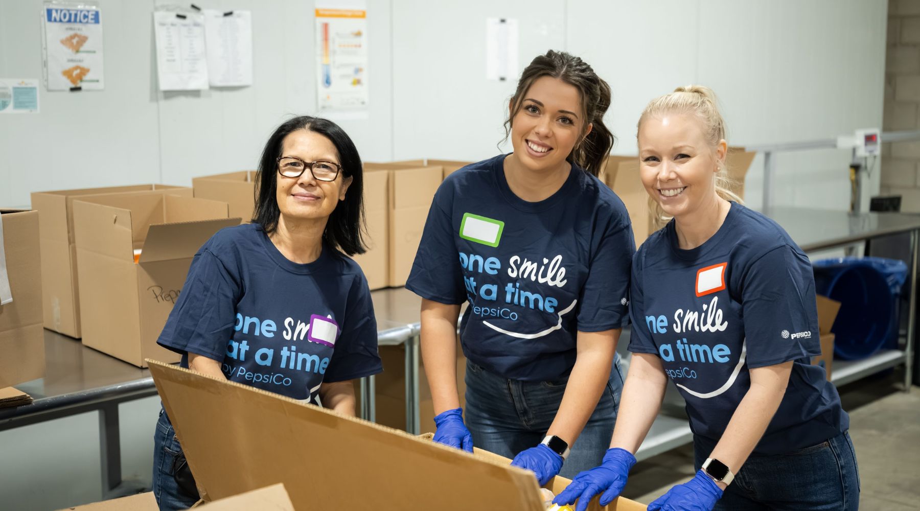 Three PepsiCo volunteers wearing gloves assemble cardboard boxes at a workstation in the warehouse.